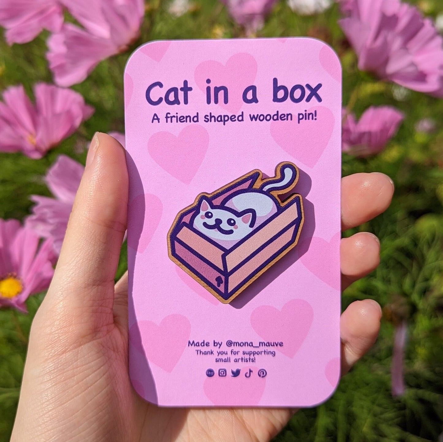 A cute wooden badge with an illustration of a smug cat in a box. The pink backing card reads: "Cat in a box! A friend-shaped wooden pin"